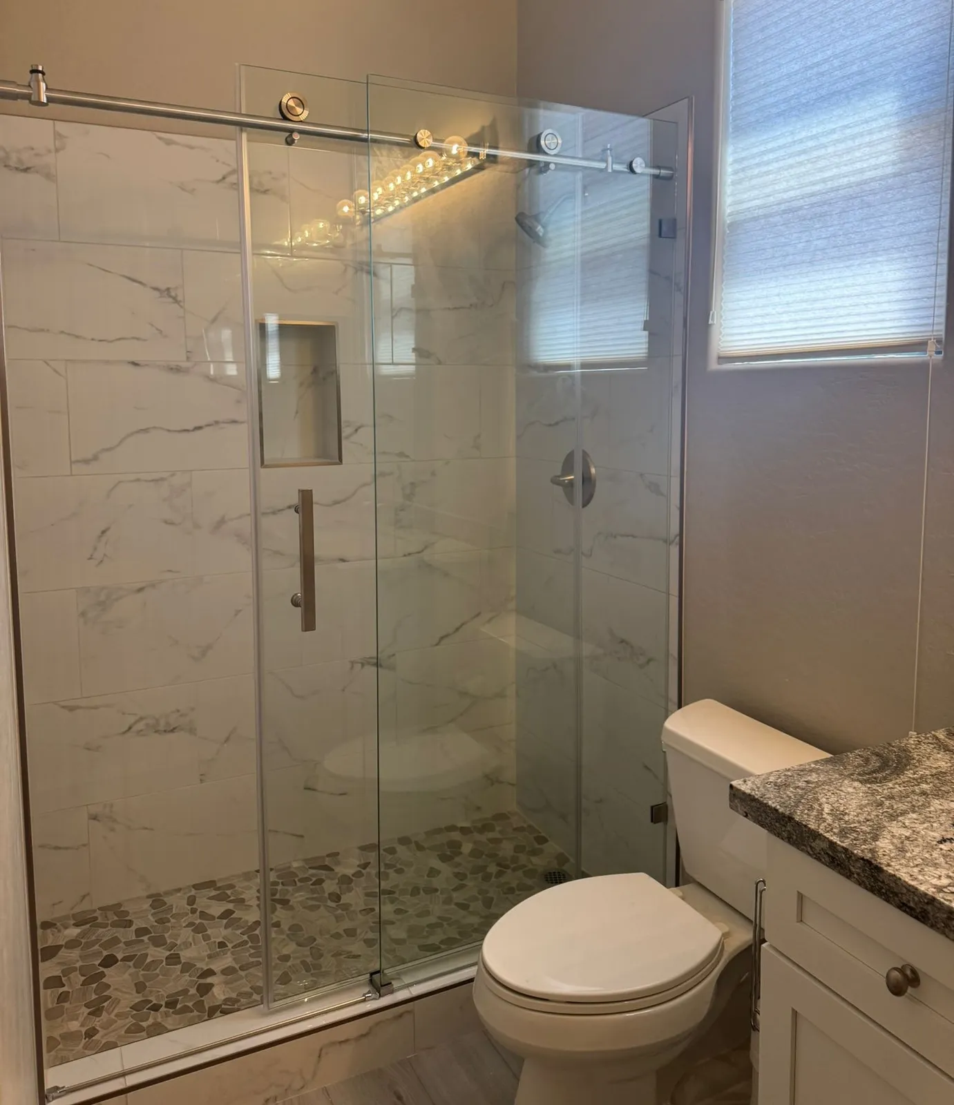 Marble-look porcelain shower with a sliding glass door, a niche and a pebble floor by RDC Renovation, serving Buckeye AZ